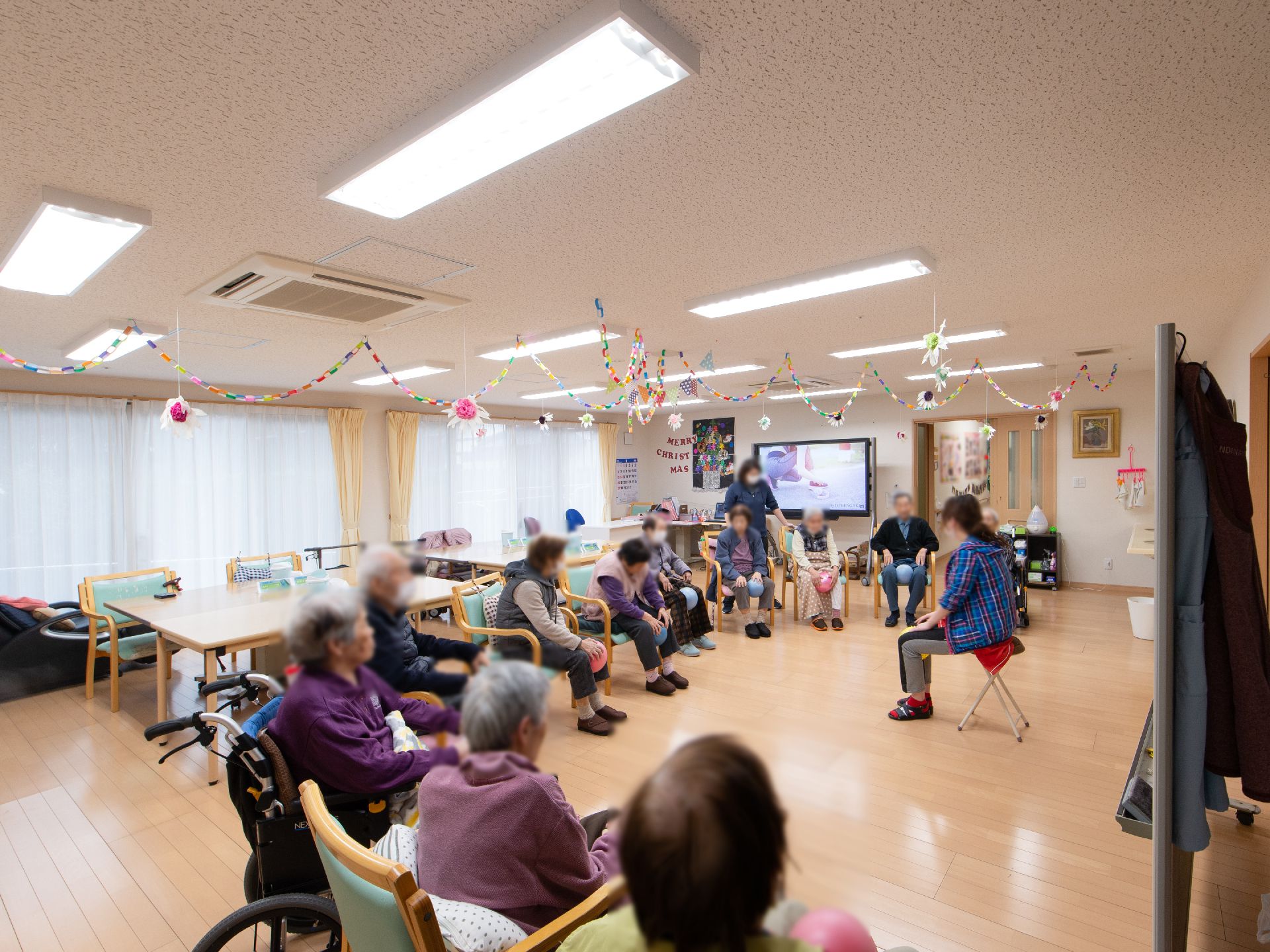 広い共有スペースに入居者様が集まって、レクリエーションをおこなっている。半円になって椅子に座り、膝にボールを挟んでいる。