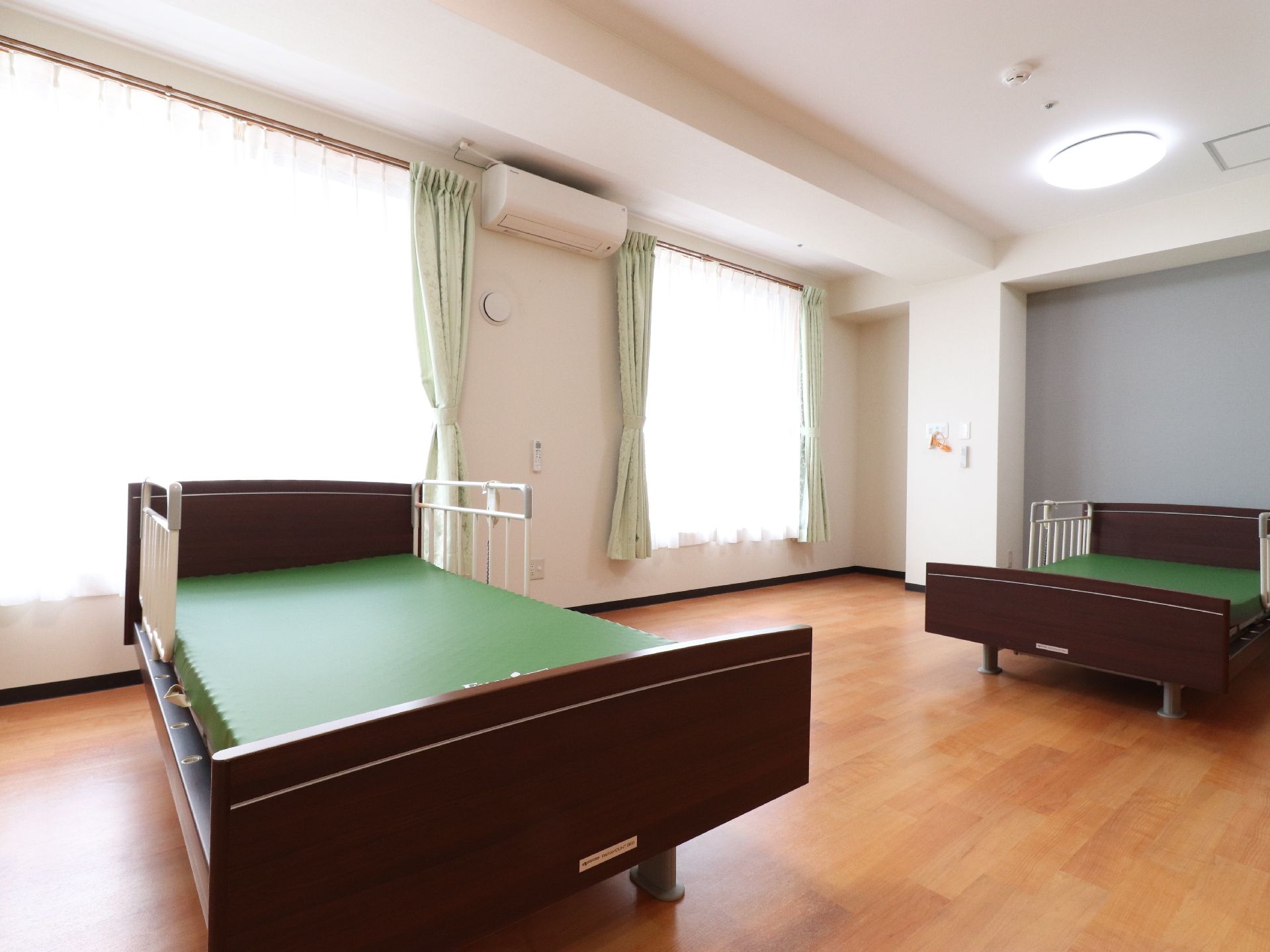 大きな窓があり、明るく開放的な部屋にはベッドが2つ用意されている。壁にはエアコン、天井には照明が設置されている。