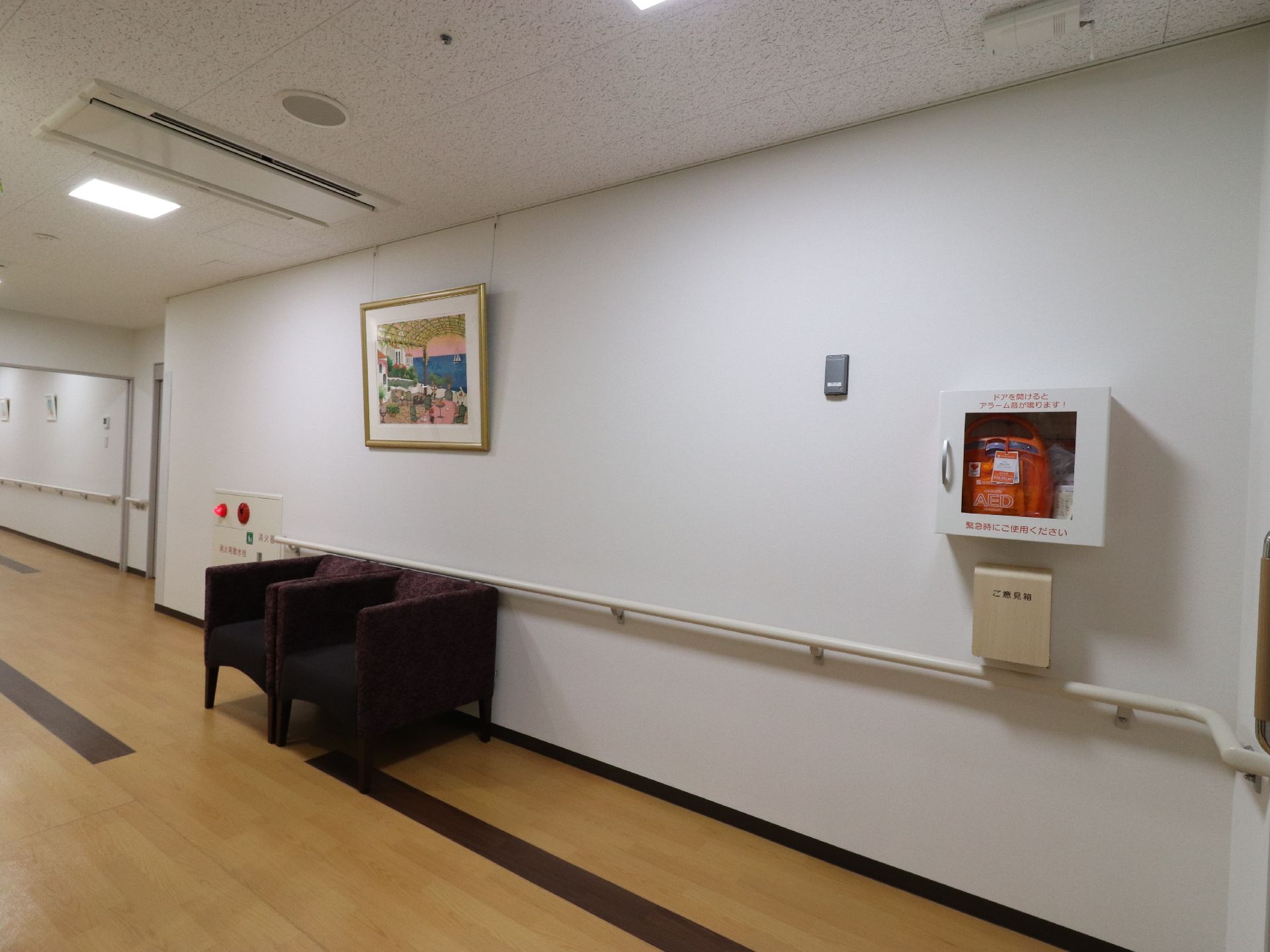 通路の幅は一定ではない。床はベージュに茶色のラインがある。壁には手すりがありAEDと絵画がある。絵画の下にソファが2脚ある。