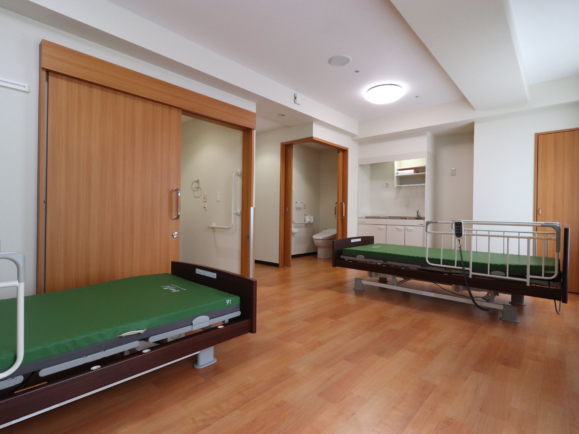 広々とした居室にはべットが2つあり、奥にはキッチンやトイレがある。天井にはシーリングライトが設置されている。