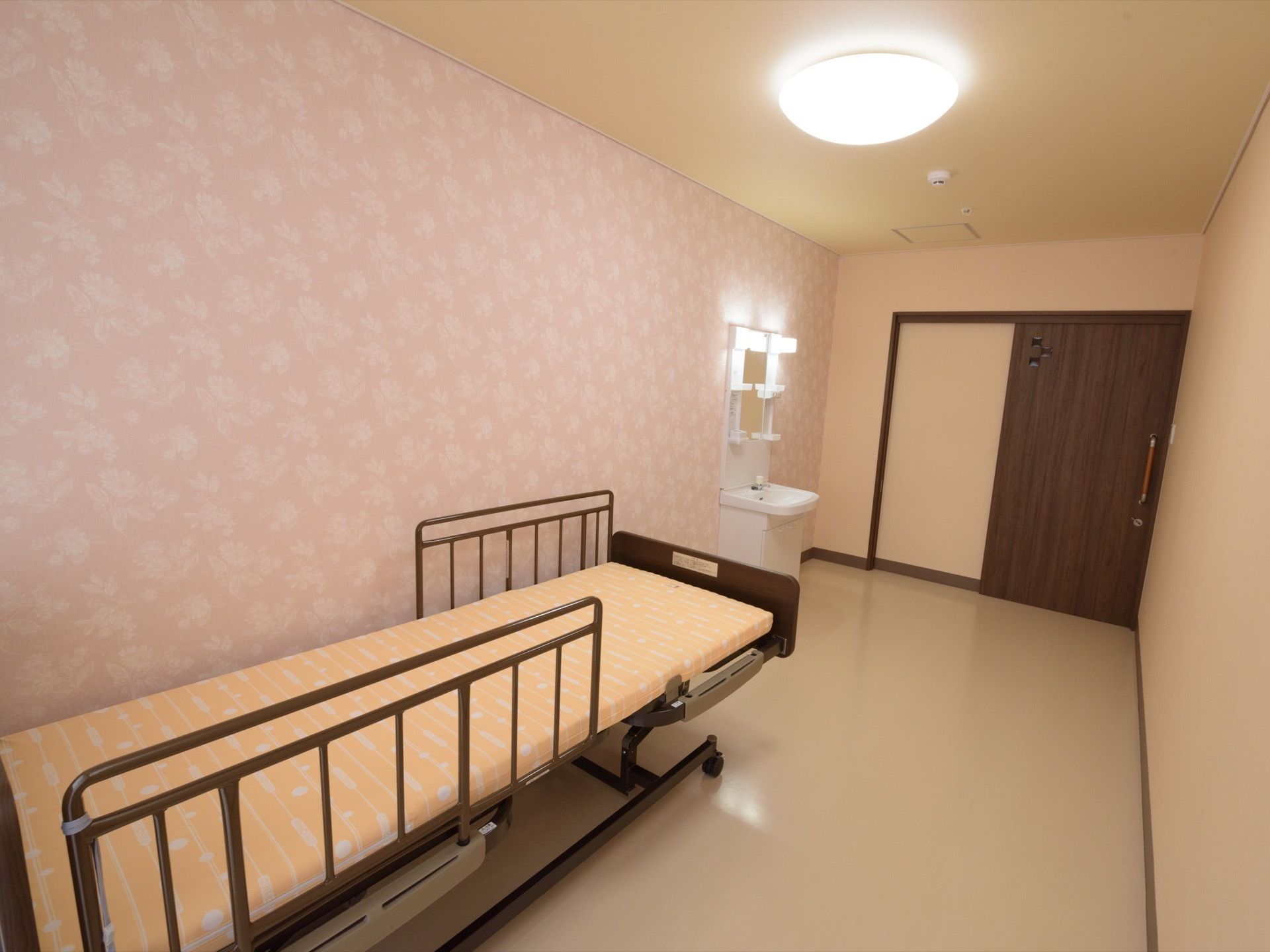 居室の壁は淡いピンク色を基調とし、白色の花柄が施されている。床はベージュ色である。ベッドには転落防止柵が備わっている。