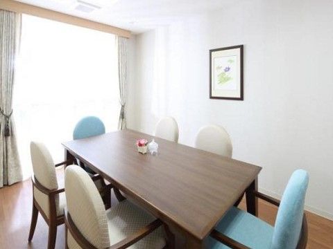 窓から明るい光が差し込む部屋の中央に、長方形の机がある。その左右には白い椅子が２脚ずつ、手前と奥には水色の椅子が置かれている。