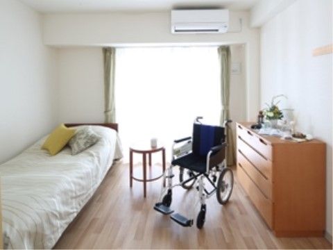 フローリングの床にはベッド、タンス、テーブルが置かれ、車椅子での移動が可能になっている。壁は白色基調で、ガラス引き戸が設置されている。