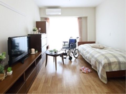 大きな窓がある明るい部屋。低床型ベッドが置かれた反対側の壁に、低いテレビ台と整理タンス、クローゼットが並ぶ。間には車椅子とテーブルも置ける。