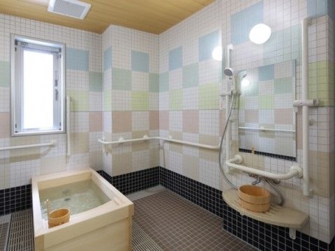 浴室には浴槽が置かれ、壁やシャワー周辺には手すりが設置されている。淡い色合いのタイルでカラフルな市松模様が描かれており、明るい雰囲気である。