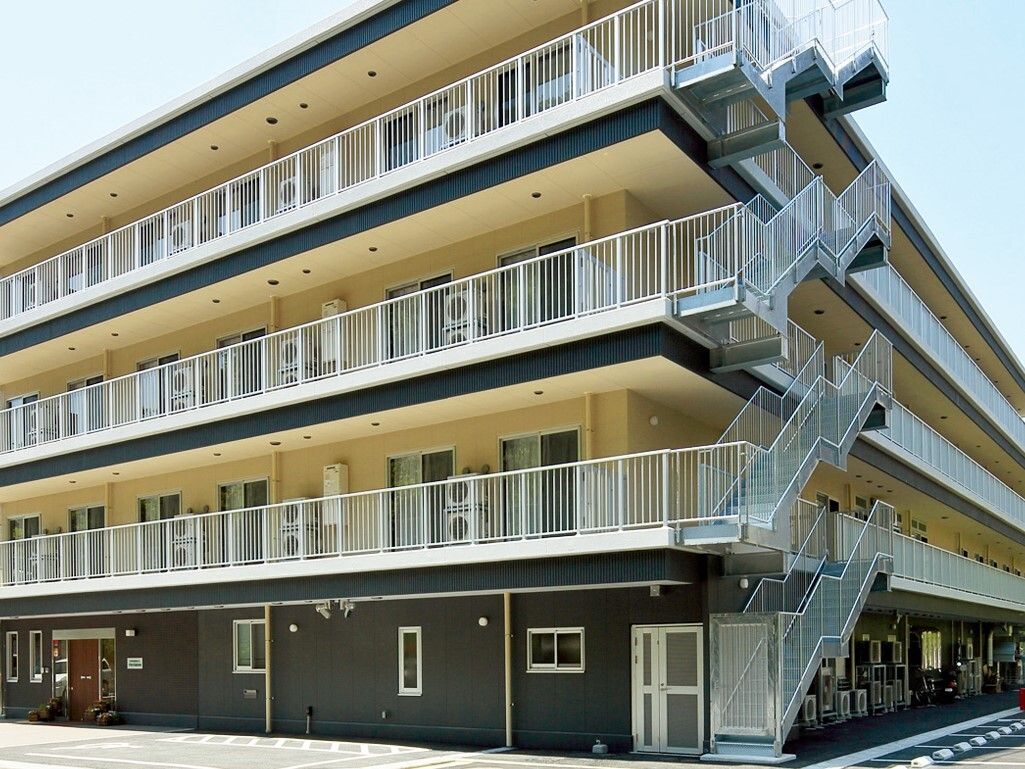 焦茶色とクリーム色が落ち着いた印象を与えるマンションタイプの施設。建物の目の前にある駐車場には、広いスペースが確保されている。