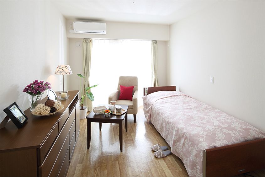 白を基調とした自然光が射し込む明るい居室である。介護用ベッドやチェスト、テーブルセット、エアコン、カーテンが備わっている。