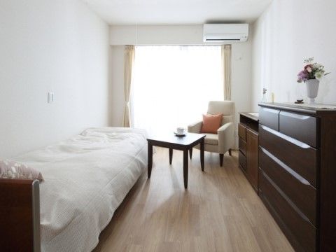 床はフローリング、白い壁に大きな窓がある明るい居室。ナースコールが備え付けられており、安心してお過ごしいただける環境である。