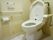 トイレには白い便座カバーと白いマットが付けられている。壁側には手すりとナースコールボタンが設置されている。