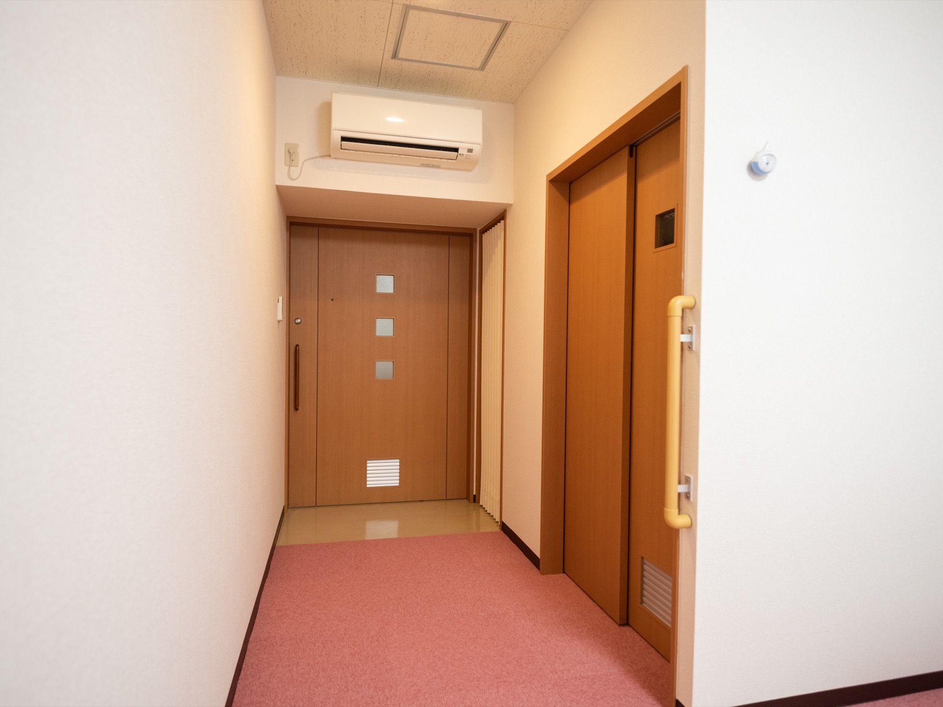 ピンクの絨毯が敷かれた居室の入口は、幅のある引き戸を採用し、入口上部にエアコンを設置。洗面スペースの扉は、片引き戸になっている。