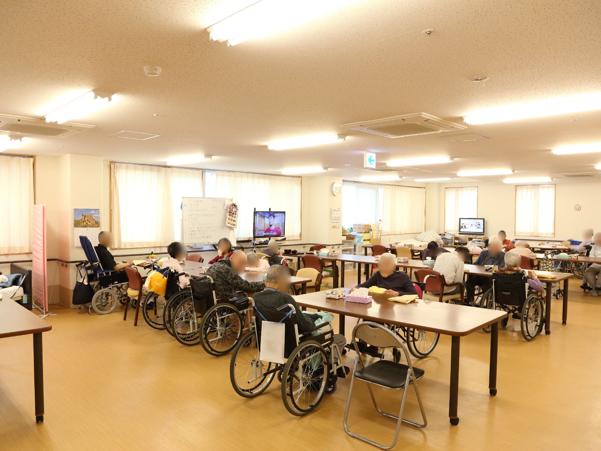 食堂には入居者様が十名ほどいる。テレビが二台あり、どちらもついている。会話をしている人もいる。窓にはカーテンがついている。