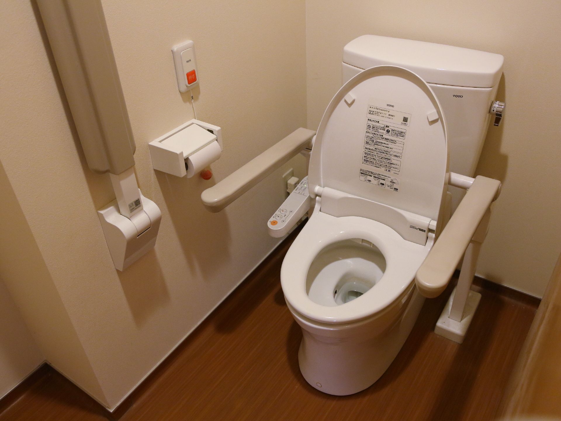 白い壁と木目調のフロアのトイレ。便器の両サイドに手すりが設置されている。壁には緊急の場合のナースコールが取り付けられている。