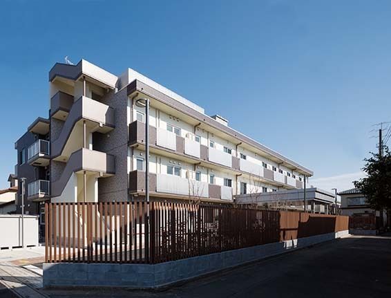 外階段があり、マンションのような建物である。道路と敷地の境に茶色い柵が設置され、内側には植物が植えられている。