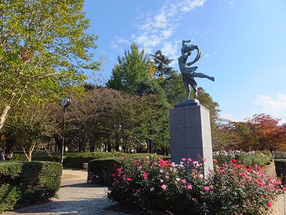 広い公園で、赤いバラがたくさん咲いている。奥の方にはテントがあり、休憩する場所も見える。緑深い大きな木が生い茂っている。
