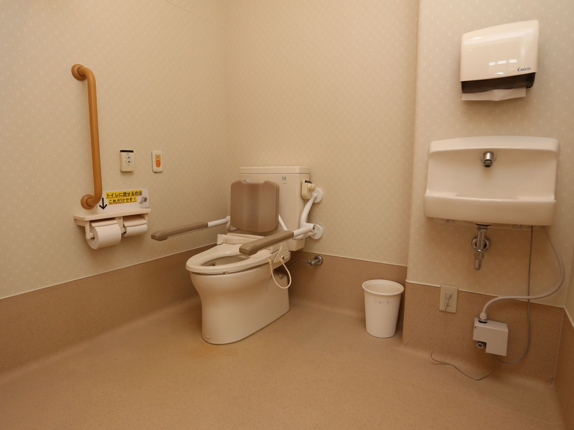 ベージュ形でまとめられた落ち着きのあるトイレである。便座には手すりと背もたれがある。手洗いとペーパータオルが設置され、非常ボタンも見える。