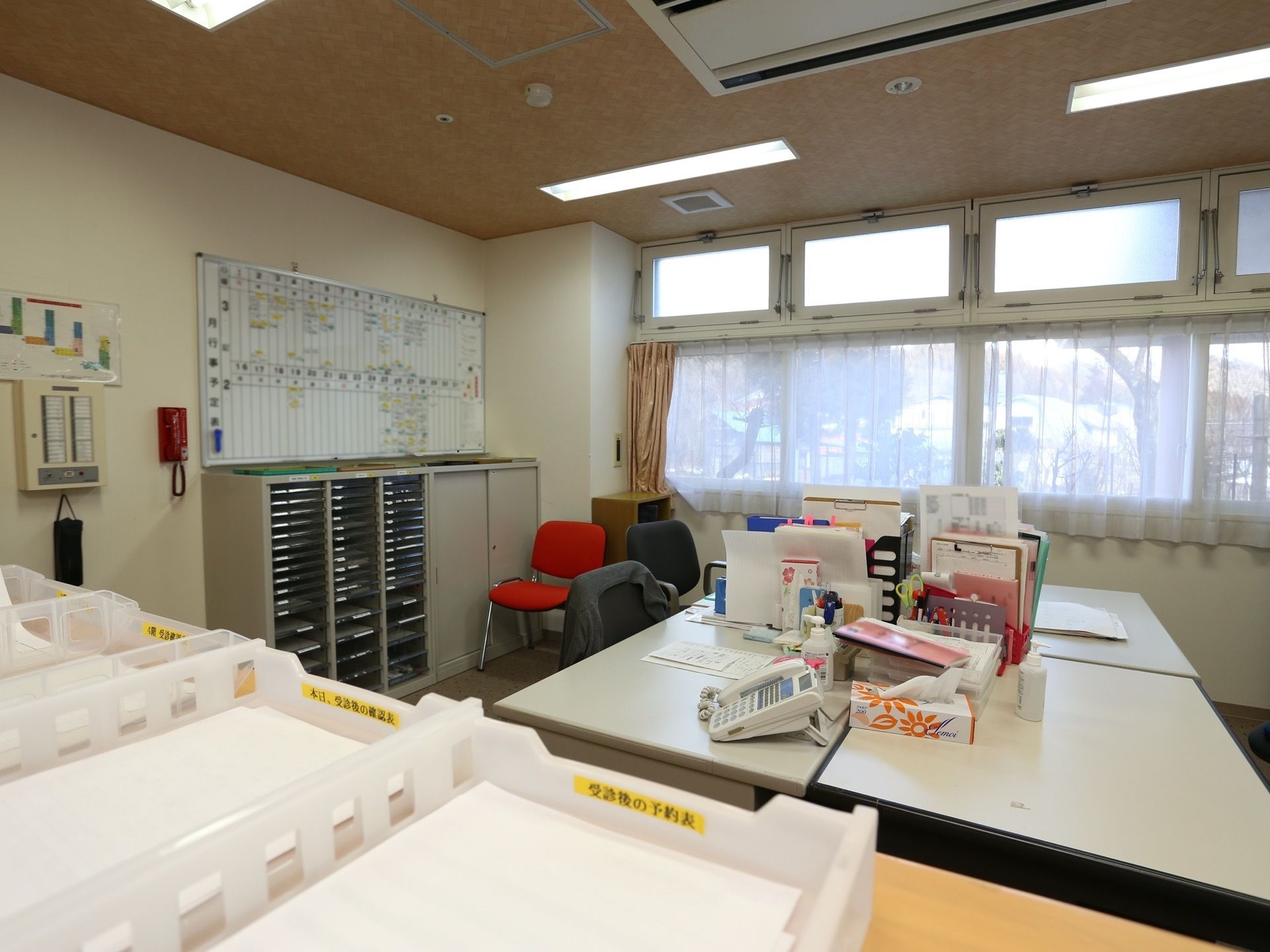 スケジュールボードや大きな書類のキャビネットが置かれたスタッフルーム。中央にデスクが4台設置してある。カウンターには書類ケースがある。