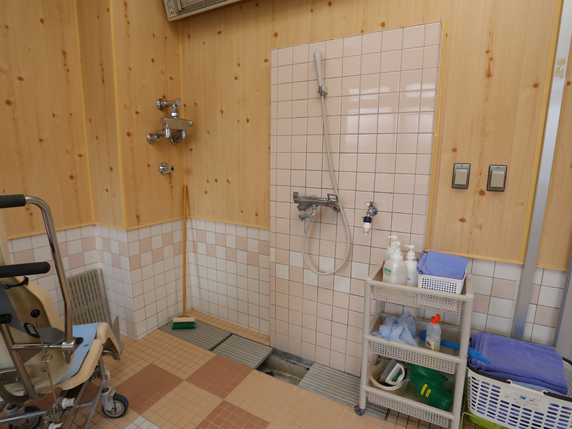 浴室の腰壁、床はタイル貼り、上部にはヒノキが使ってある。介護用のシャワー椅子とデッキブラシや洗剤等があり、清掃中である様子がうかがえる。