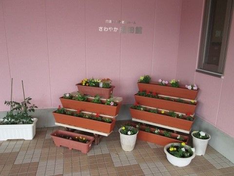 紫色の外壁で施設名が書かれている。角地にはプランターが段になった台に並べられ、可愛らしい花が咲いている。