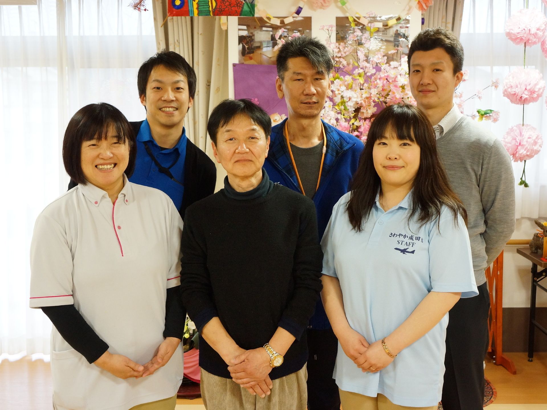 様々な年代の、様々な制服を着たスタッフが6名並んでいる。後ろの壁には写真が貼ってあり、干支の置物や花などが飾ってある。