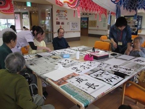テーブルには新聞紙が敷かれ、入居者様が習字を行っている。空いているスペースに「成長」「さわやか」等書かれた半紙が置いてある。