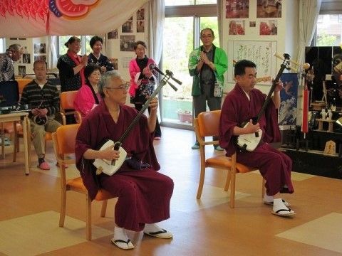 紅色の着物を着た男性二人が、三味線で演奏をしている。後ろには法被を着た男性と女性が立っている。天井には巨大な鯉のぼりが吊るされている。