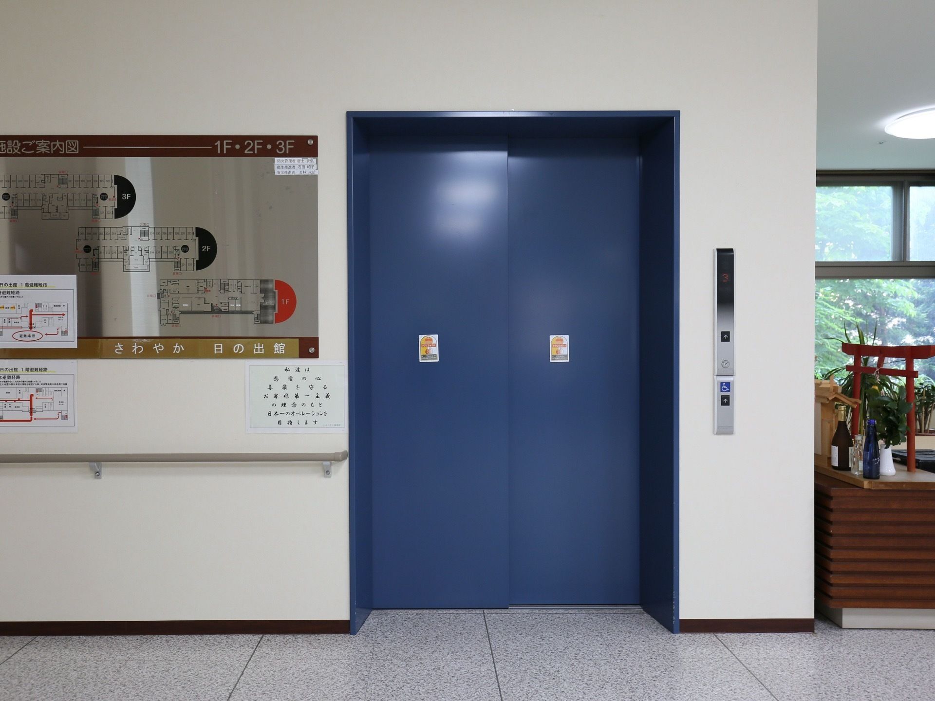 玄関ホールにエレベーターの乗降口がある。左側の壁に、フロアごとの案内図が表示されている。その下に手すりが設置されている。