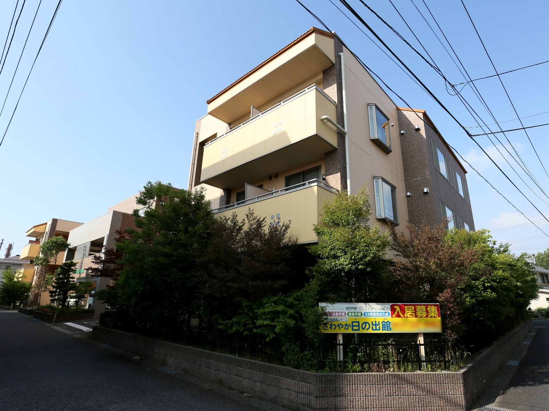 ２面が道路に面して建っている３階建ての施設である。敷地角部の植栽の前に、入居者様を募集する看板が設置されている。