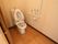 取っ手がついた引き戸を開けるとトイレがある。壁には大きなＬ字型の手すりが設置されている。オレンジ色の緊急呼び出しボタンがついている。