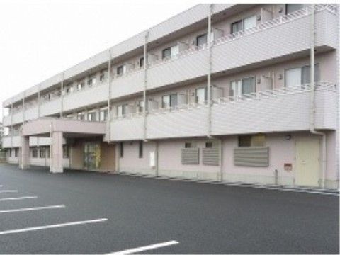 ３階建ての白を基調とした大きなグループホーム。屋根付きの玄関で、建物前には広い駐車場が設けられている。