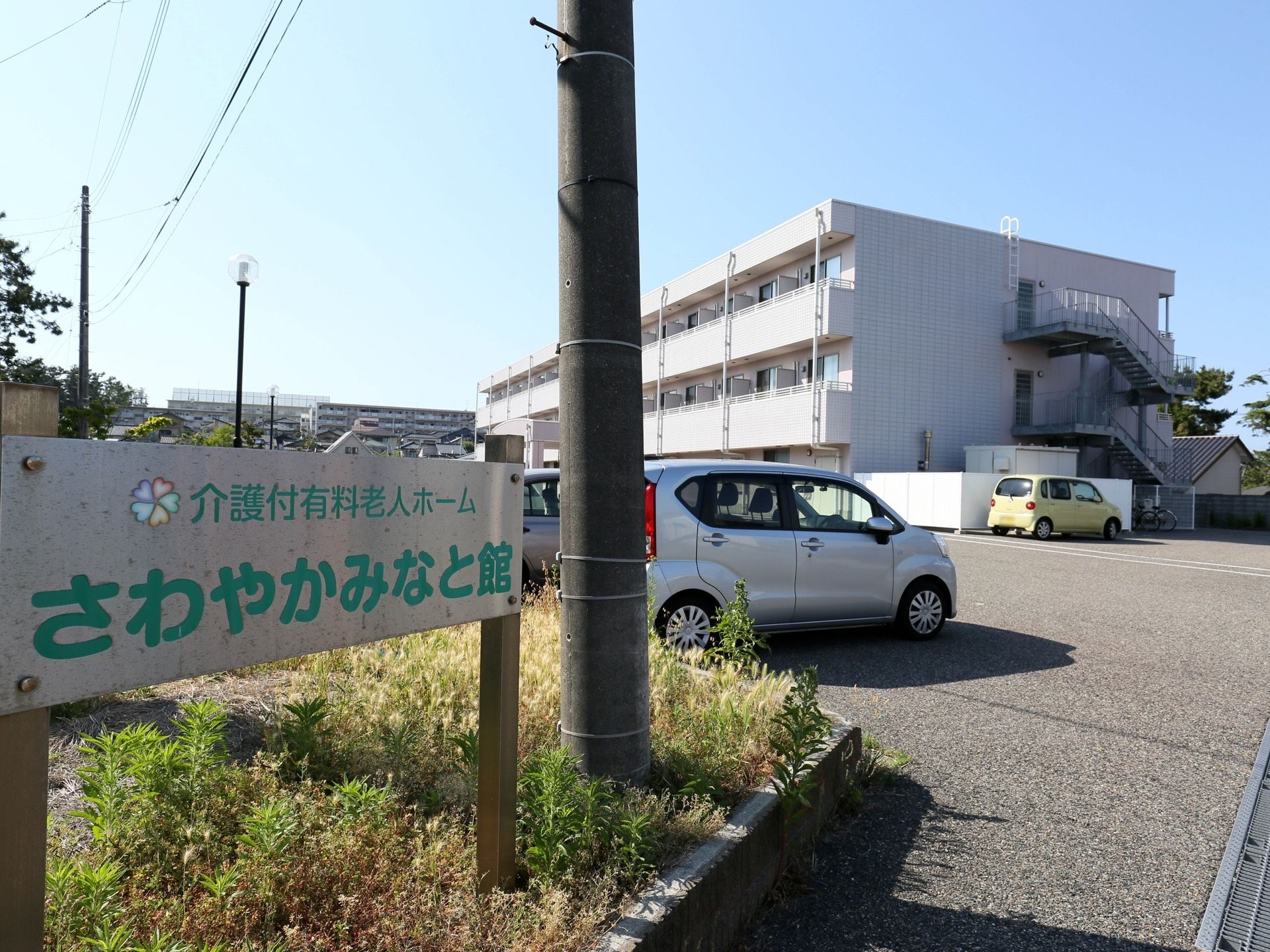 敷地入口に、銀色のプレートに緑色の大きな文字で施設名称が書かれた看板が設置されている。その横には電柱が立っている。