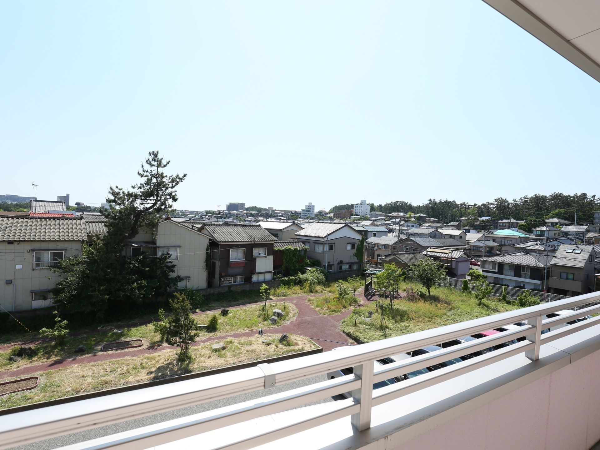 居室のバルコニーから周囲の家並みが見える。敷地内には遊歩道が整備された庭園があり、背の高い木が植えられている。
