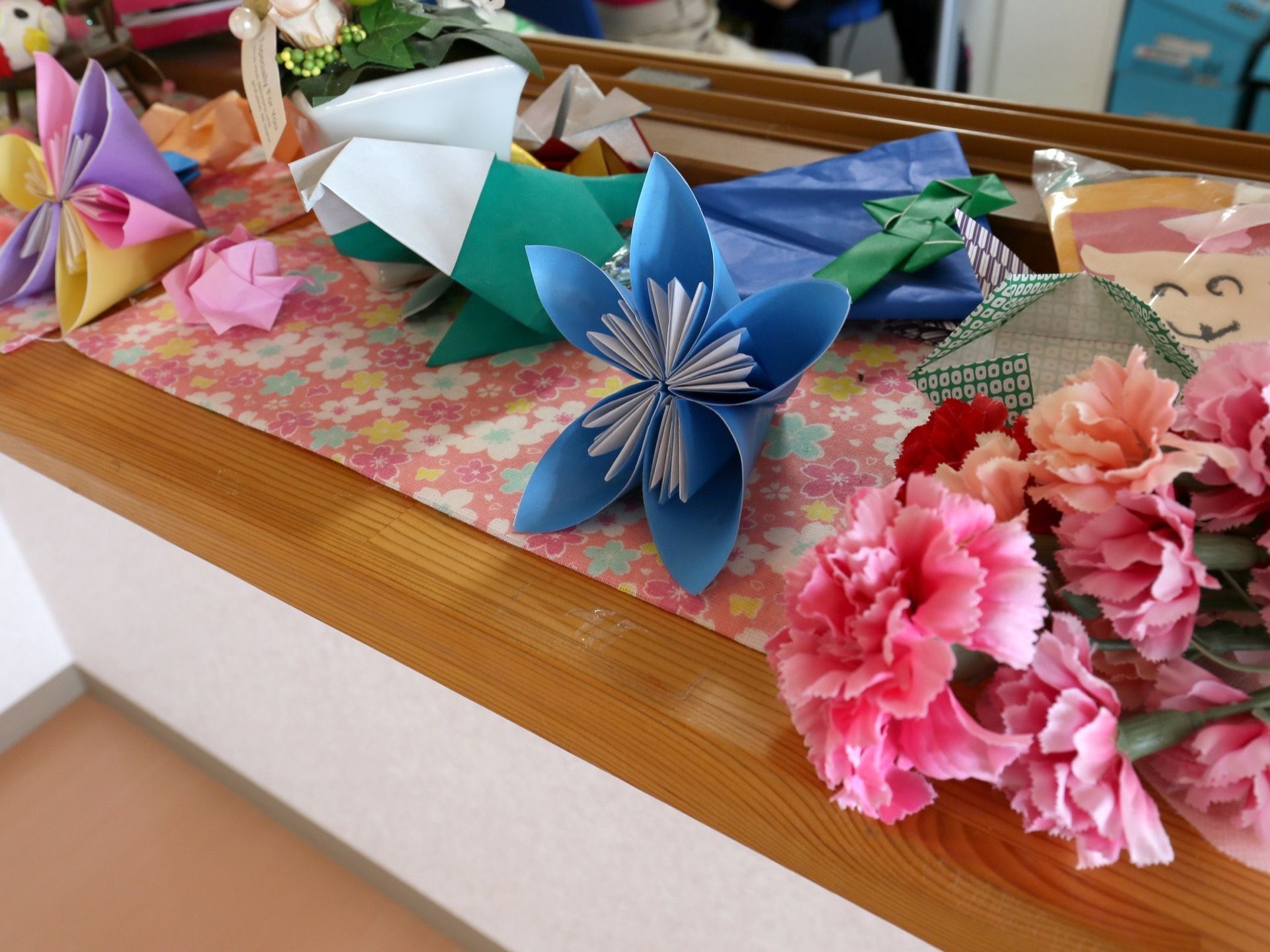 入居者様が色とりどりの紙で制作した作品が、棚板の上にたくさん飾られている。下には花柄のマットが敷いてある。