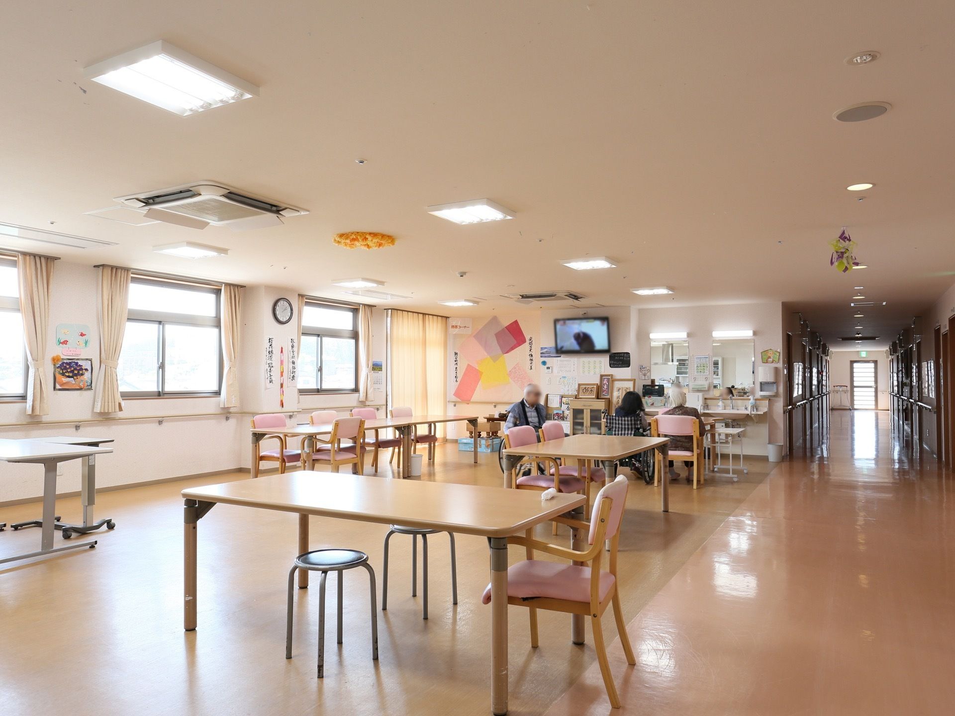 入居者様が３名、食堂のテーブルについている。壁掛けテレビが設置されていて、その右側には洗面台が２台ある。