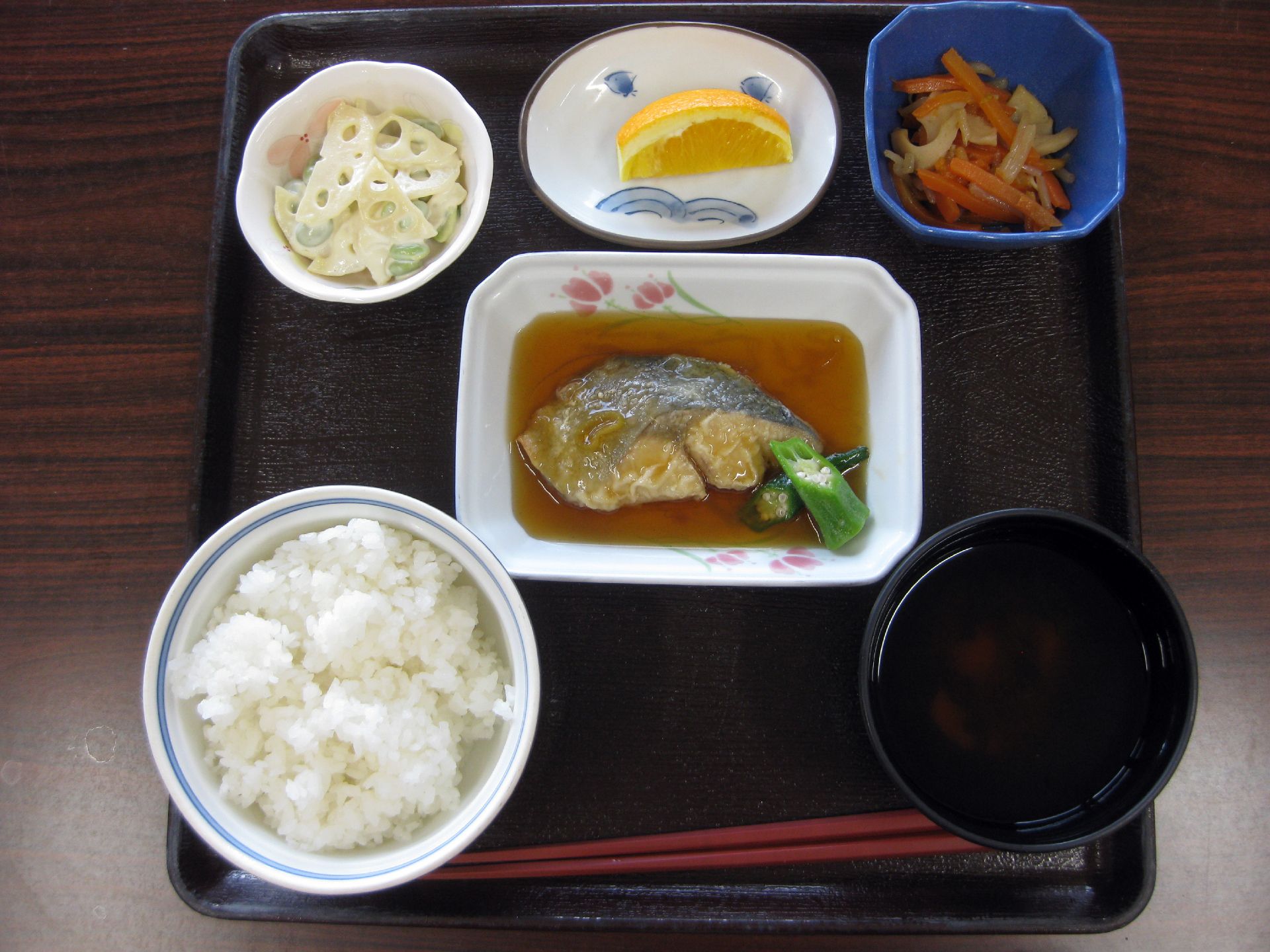 メインの魚料理、ニンジンやもやしなどが入った野菜炒め、レンコンサラダ、レモン、ご飯と汁物が、黒いトレーに並べられている。
