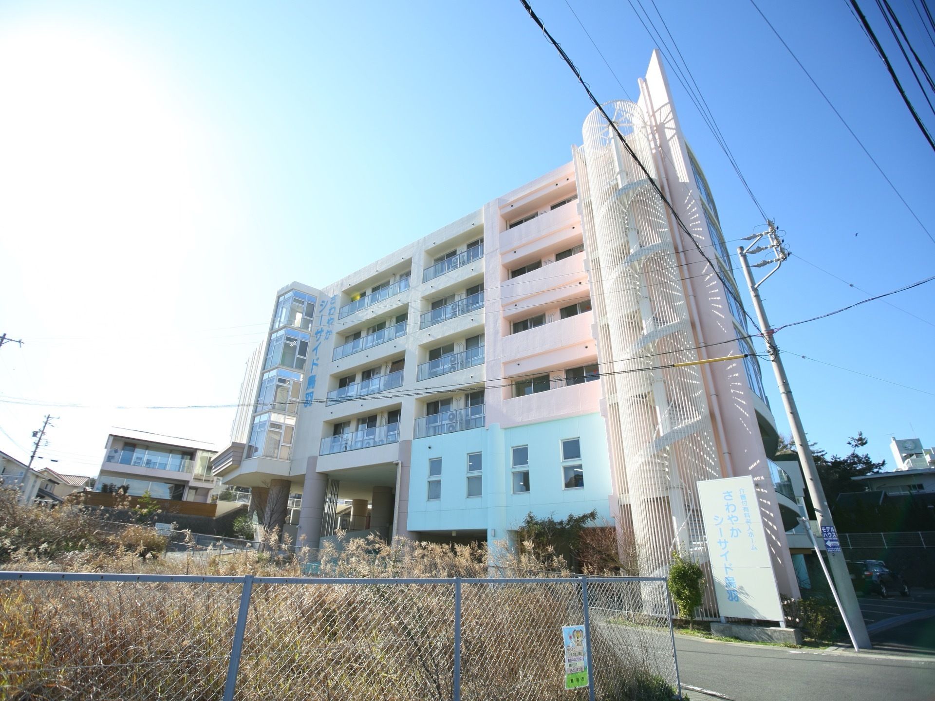 建物の道路側に、螺旋状の外階段が設置されている。外壁には、大きな文字で施設名が縦に書かれている。居室のところには、ベランダがある。