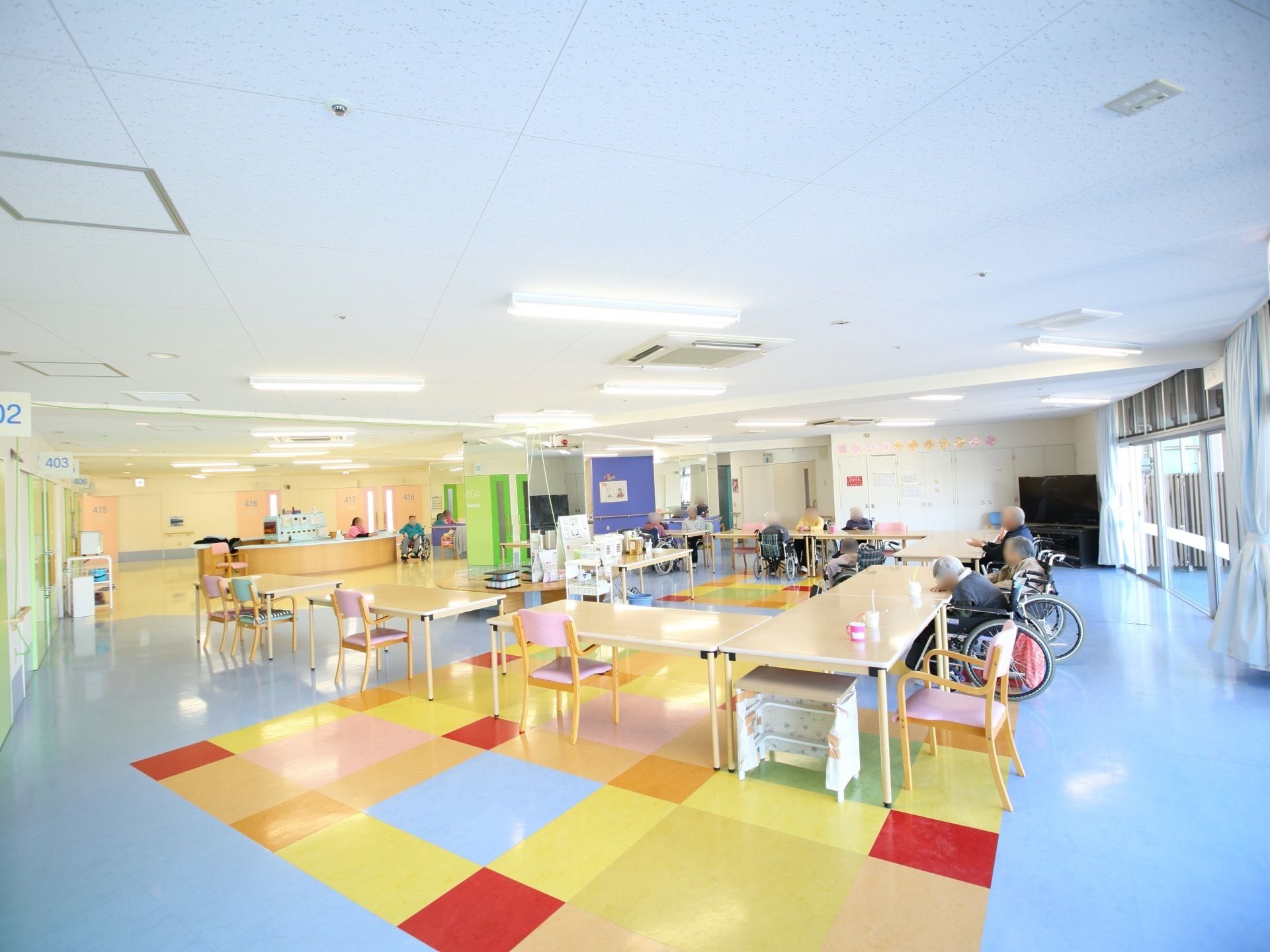 広いリビングで、テーブルがコの字型に置かれている。車椅子に乗った入居者様が、窓の側のテーブルに3人いる。