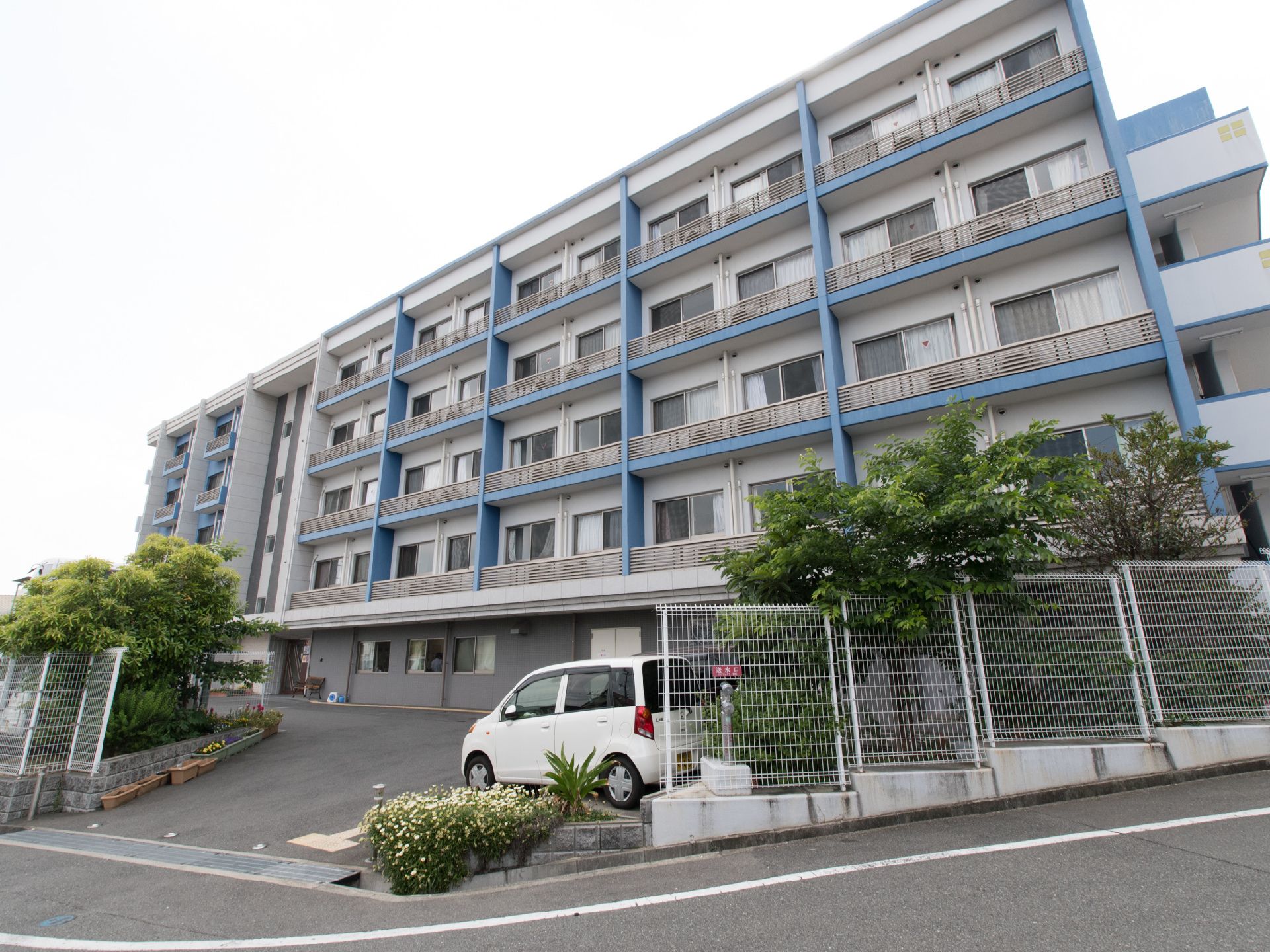 建物の前から道路へと続く道は、なだらかな坂道になっていて、その周辺に草木や花が植えてある。入口付近には車が置いてある。