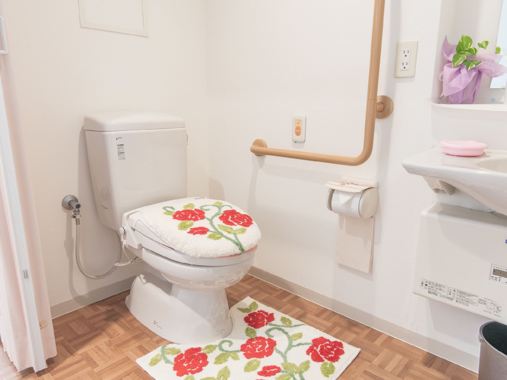 白い壁に、ベージュ色のL字型の手すりが付いている。赤い花の絵柄のトイレカバーとマットが置いてあり、明るい雰囲気である。
