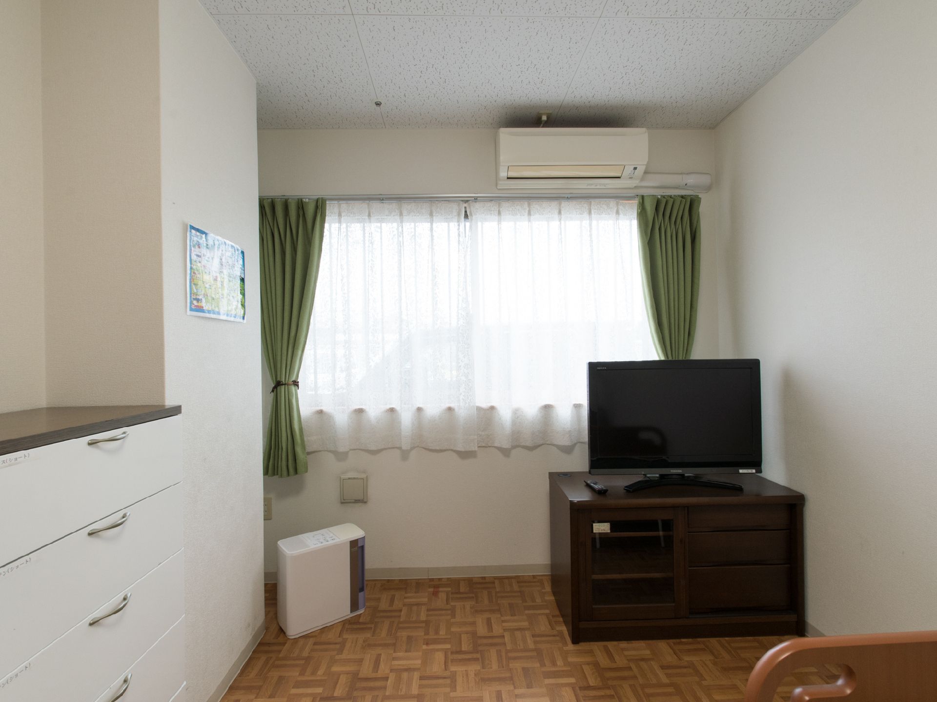 こげ茶色のテレビボードと薄型のテレビが窓際に置かれている。窓の上にはエアコンも設置されている。横の壁には、白いチェストもある。