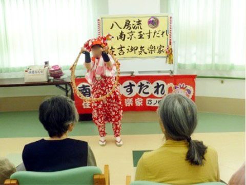 芸を披露している様子である。後ろには芸の名前が書かれた赤い横断幕と黄色い看板がある。入居者様が椅子に座って芸を見ている。