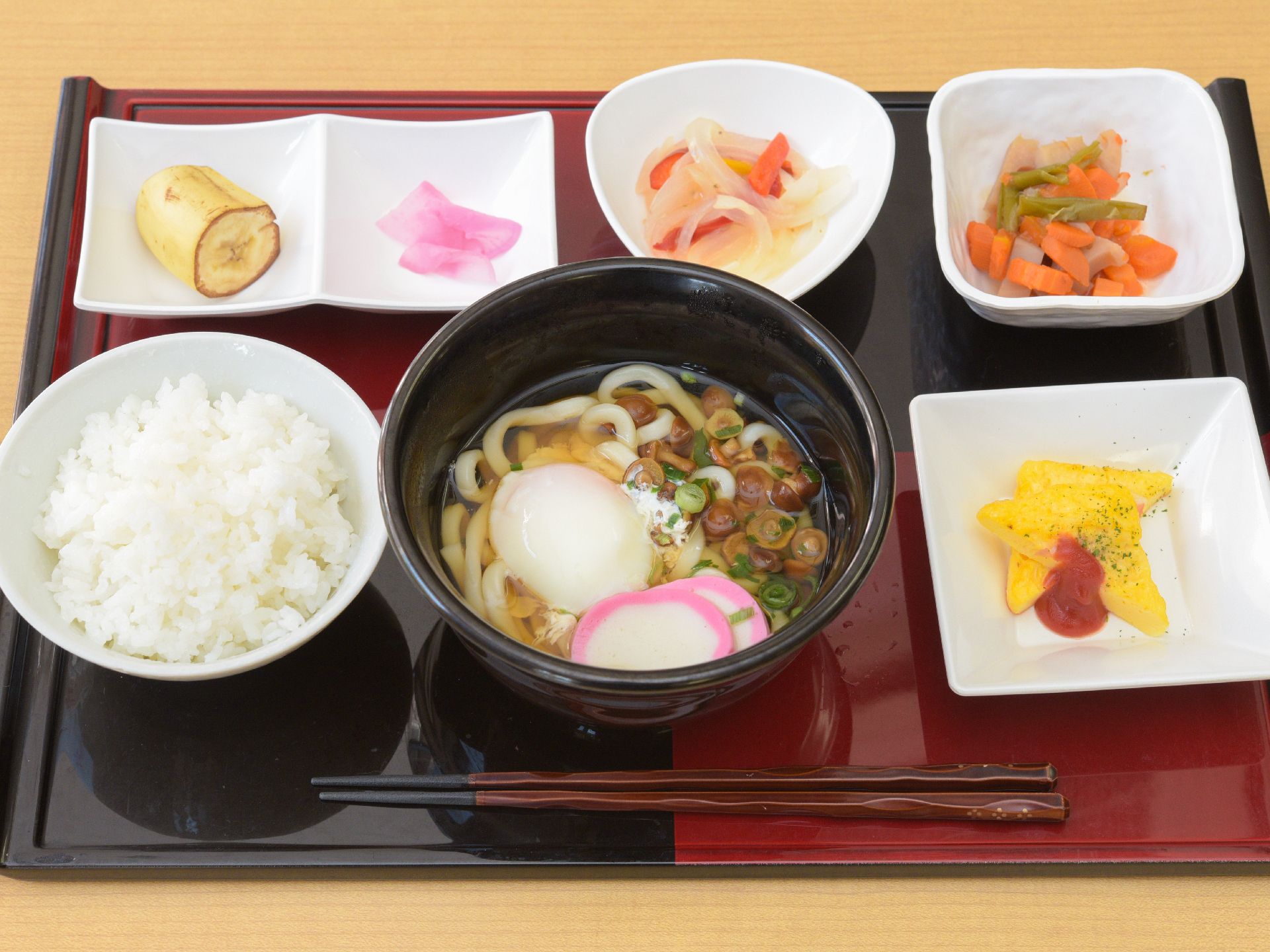 ご飯とうどん、ケチャップがかけられたオムレツ、レンコンや人参などの野菜料理、デザートのバナナなどがトレーに並んでいる。