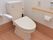 白い壁と木目調の床が落ち着いた雰囲気のトイレである。跳ね上げ式の白い手すりや、ベージュ色の横長の手すりが設置されている。