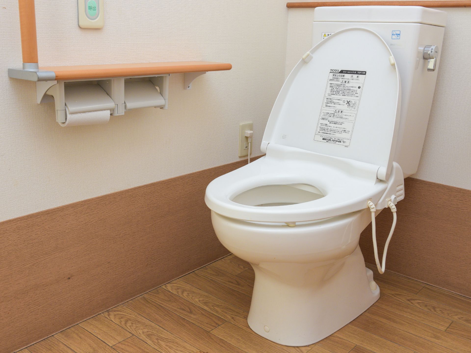 白い壁とフローリングの室内には、タンク付きのトイレが1つ設置されている。トイレットペーパーを取りつける場所が2ヶ所設けられている。