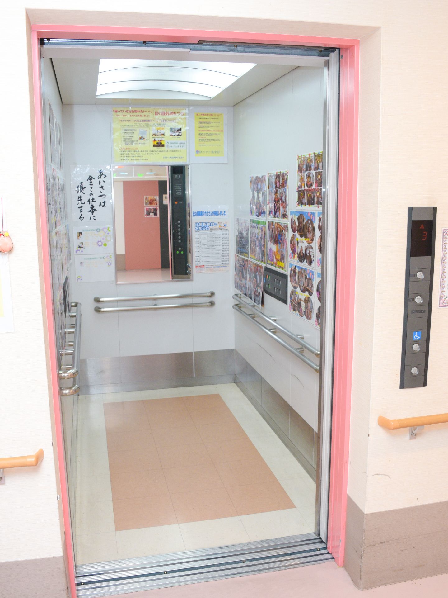ピンク色の縁のエレベーターは、奥の壁に鏡が取りつけられている。両サイドの壁には、写真が飾られている。