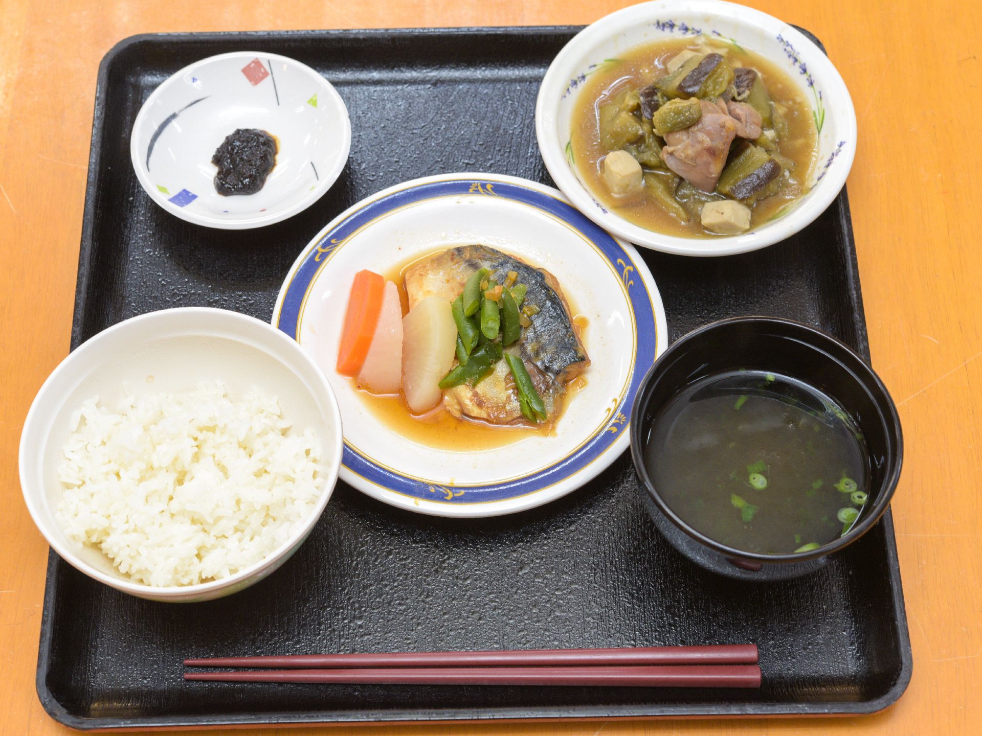黒いトレーの上には、煮魚を中心に全部で5種類の食事が並べられている。手前には、あずき色の箸が置かれている。