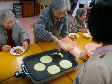 テーブル席に入居者様たちが集まっている。ホットプレートには５枚のホットケーキが焼かれており、1人の入居者様がひっくり返そうとしている。