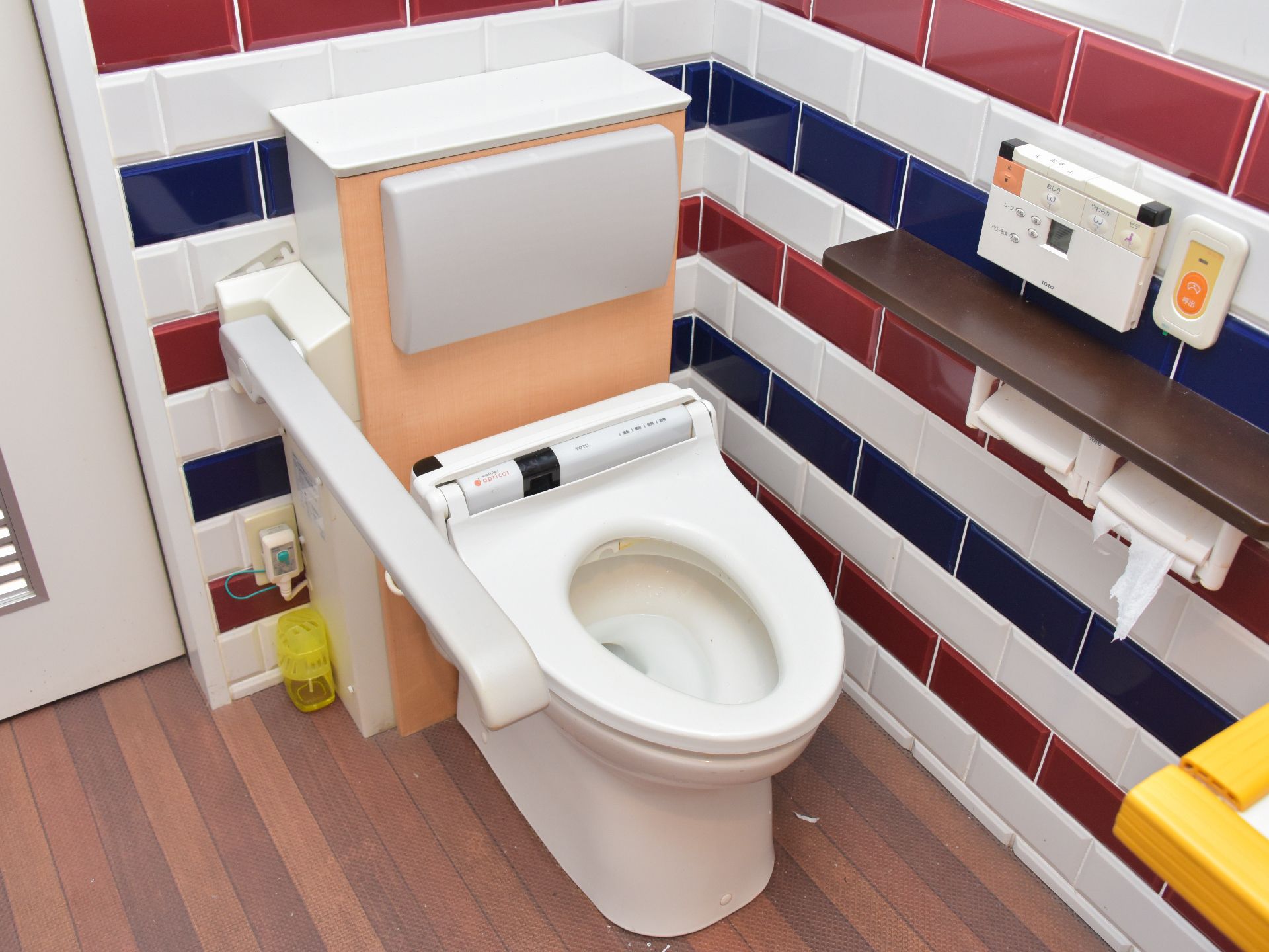 トリコロールカラーの明るいトイレである。手すり、肘置き、ナースコール、ウォシュレット、背もたれ付きと完備されている。