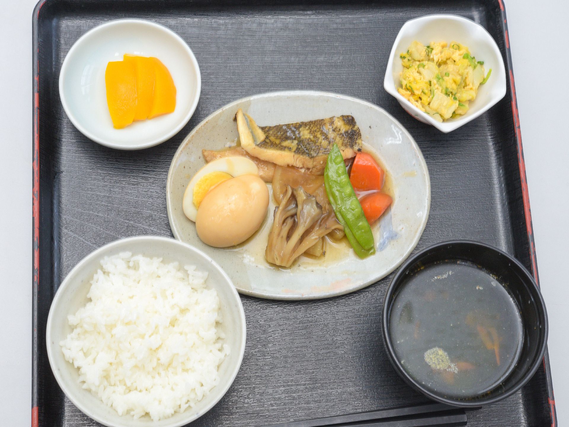 お盆に盛りつけられた、色がきれいな食事。主食のごはんを始めとして、汁物、魚の煮物、サラダ、沢庵など。