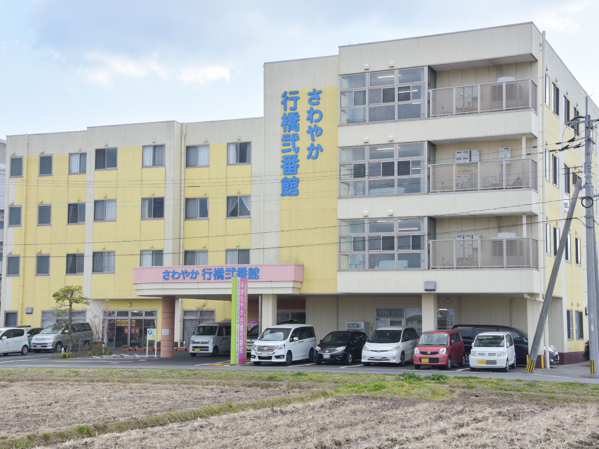 ピンクと黄色のパステルカラーの4階建て施設である。分かりやすく施設名がでており、広く駐車場が取られていて、多く車が収容できる。