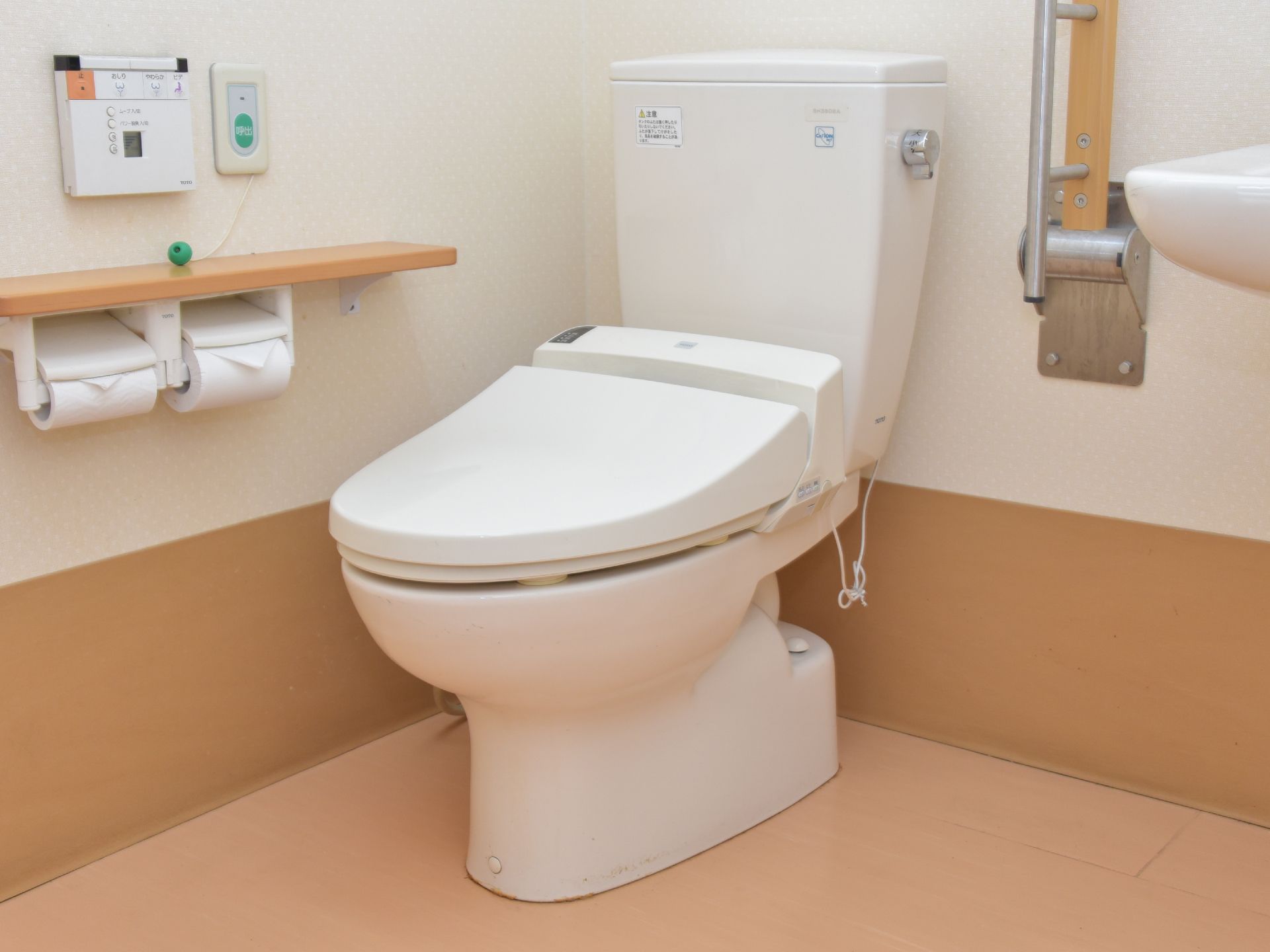 手すりのついた洋式トイレ。トイレットペーパーホルダーの上にはシャワートイレの操作パネルと呼び出しボタンが並んで設置されている。